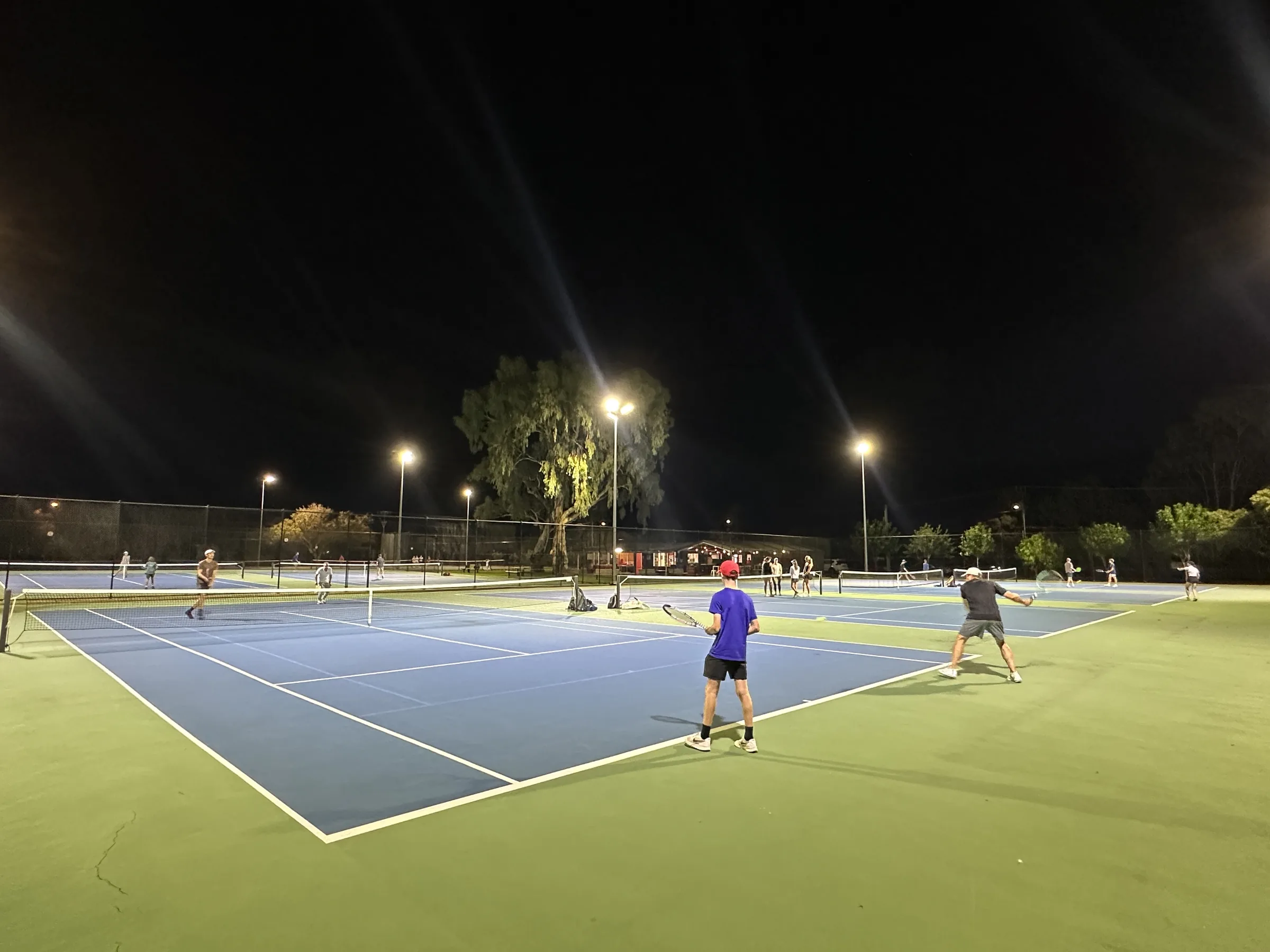 Adults playing social tennis across the courts at night